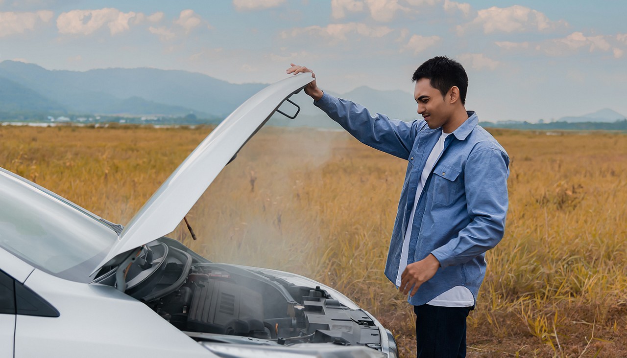 A young man on a rural road lifting up his car hood to find steam coming out of the engine area.