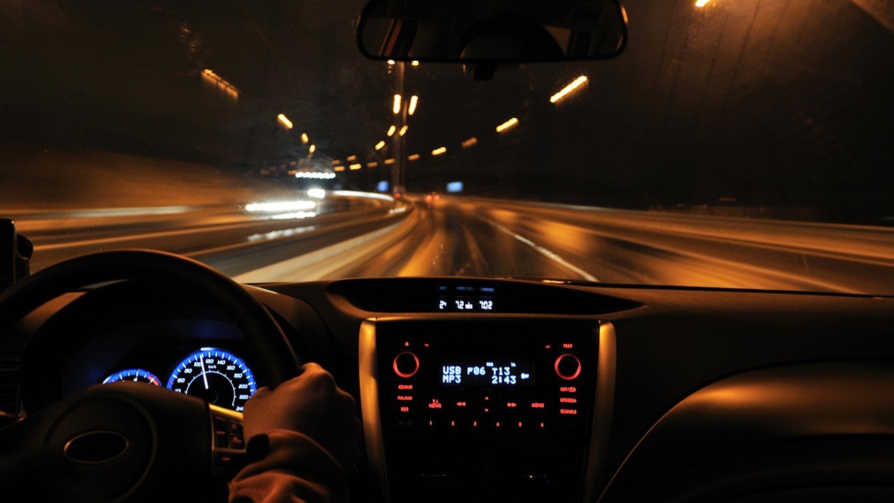 The inside of a car at night with the dash illuminated and streetlights visible in the background.