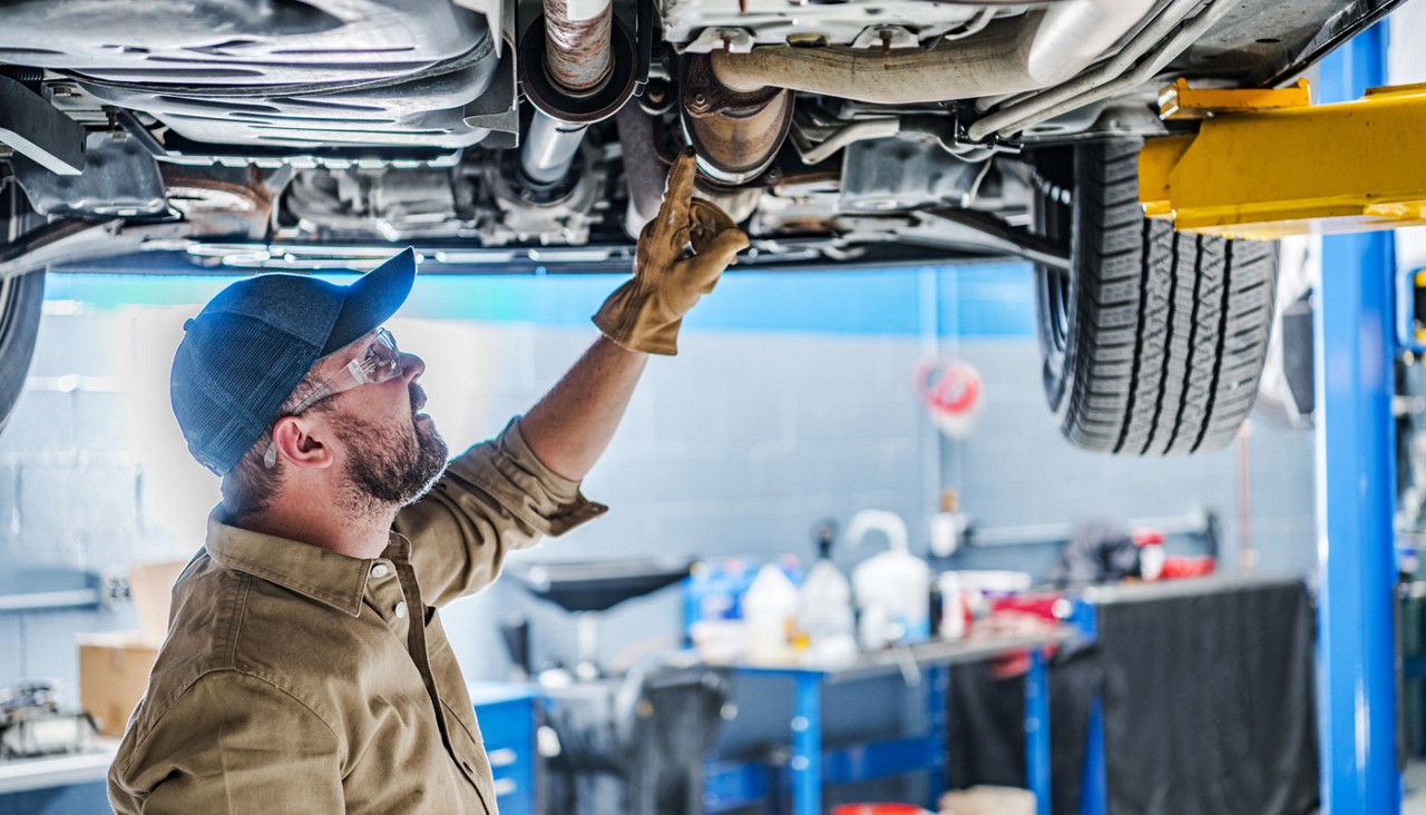 A mechanic in a ball cap looking at a catalytic converter underneath a car.