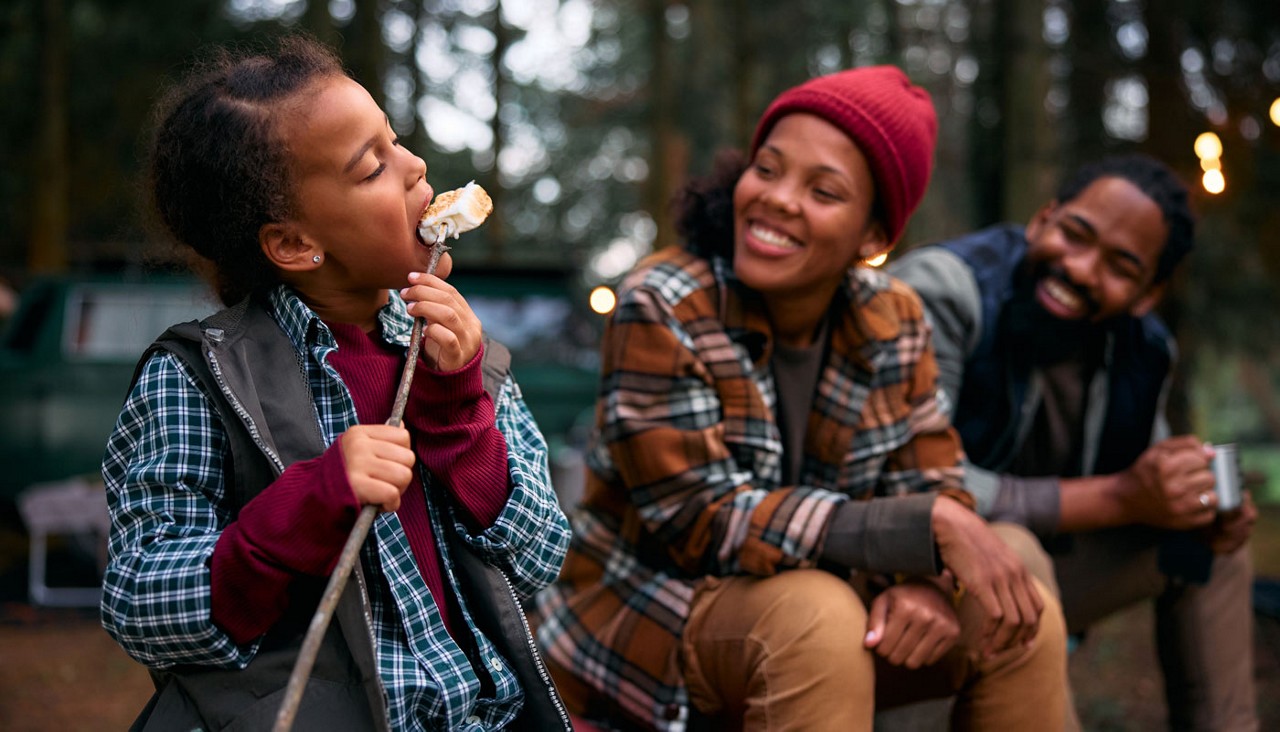 Black little girl eating roasted marshmallows while camping with parents in woods.