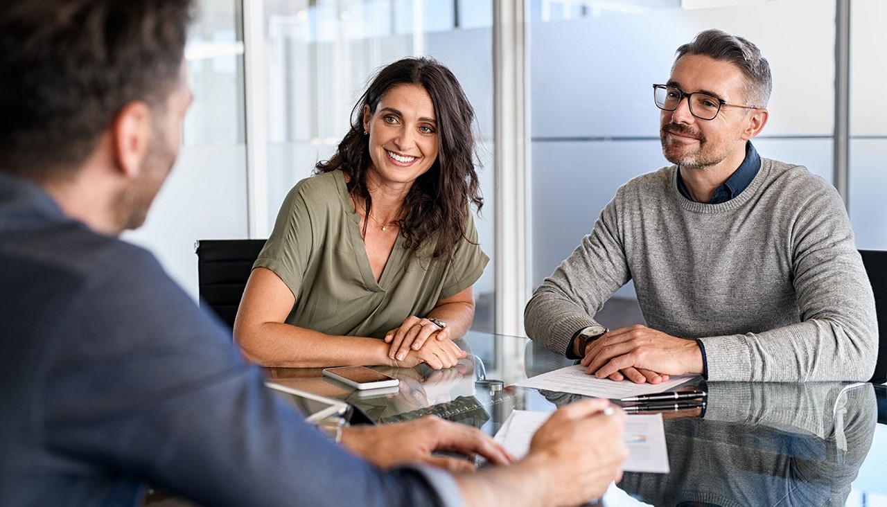 A couple sitting at a desk with paperwork while a man in a suit explains something to them.