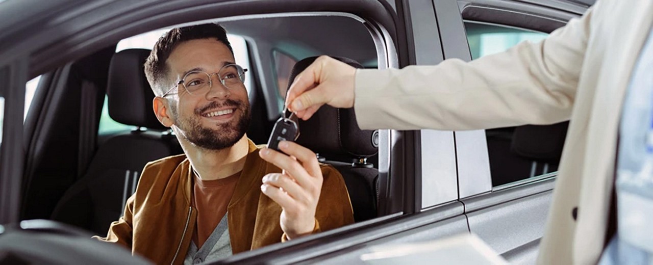 Man with dark hair and a short beard wearing a burgundy shirt sitting in the driver's seat of a car while a person in a beige coat and light colored shirt hands him the keys to his rental car.