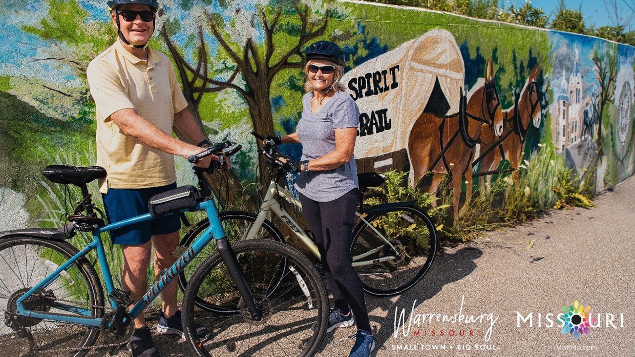 A man in black shorts, yellow polo shirt, tennis shoes, sunglasses and a bike helmet and a dark haired woman in black leggings, gray t-shirtl, tennis shoes, sunglasses and a bike helmet standing next to a wall with a mural showing a covered wagon being pulled by horses and trees.